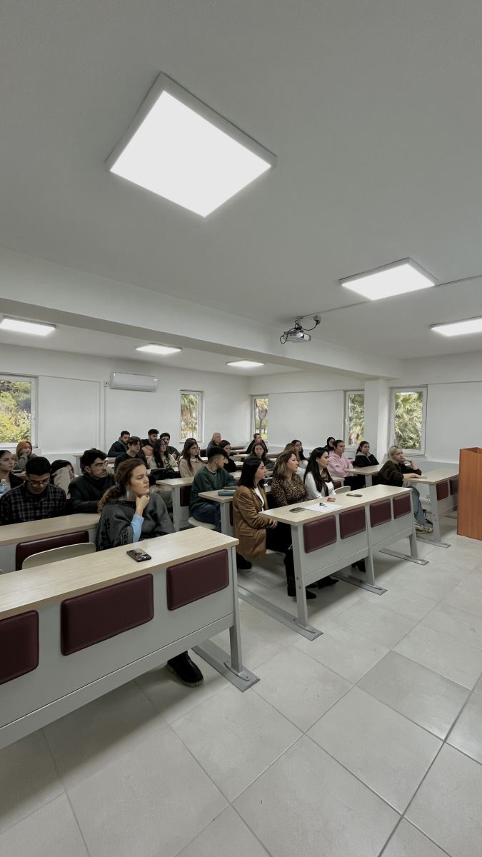 Bir sınıfta öğrenciler sıralara oturmuş, dikkatle karşıya doğru bakıyorlar. Sınıf geniş ve aydınlık; tavanda kare LED ışıklar ve bir projeksiyon cihazı bulunuyor. Öğrencilerin bazıları defterlerine eğilmiş, bazıları öğretmeni izliyor. Pencerelerden dışarıda ağaçlar ve yeşil alanlar görülüyor.